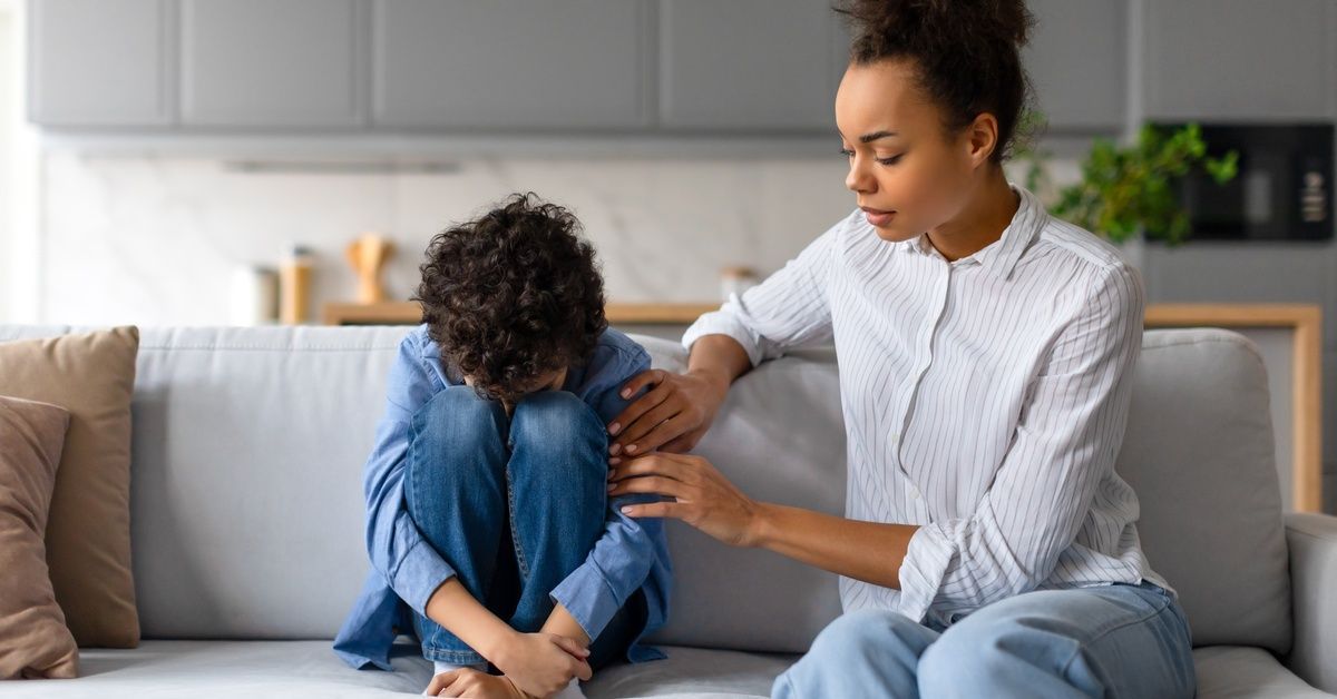A mother consoles her child on a gray sofa in their home. The child hunches over and hides his face.