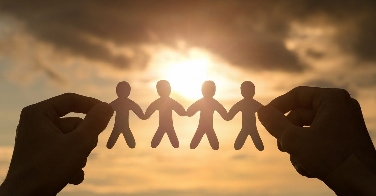 Hands hold a chain of paper people silhouetted against a glowing sunset sky.