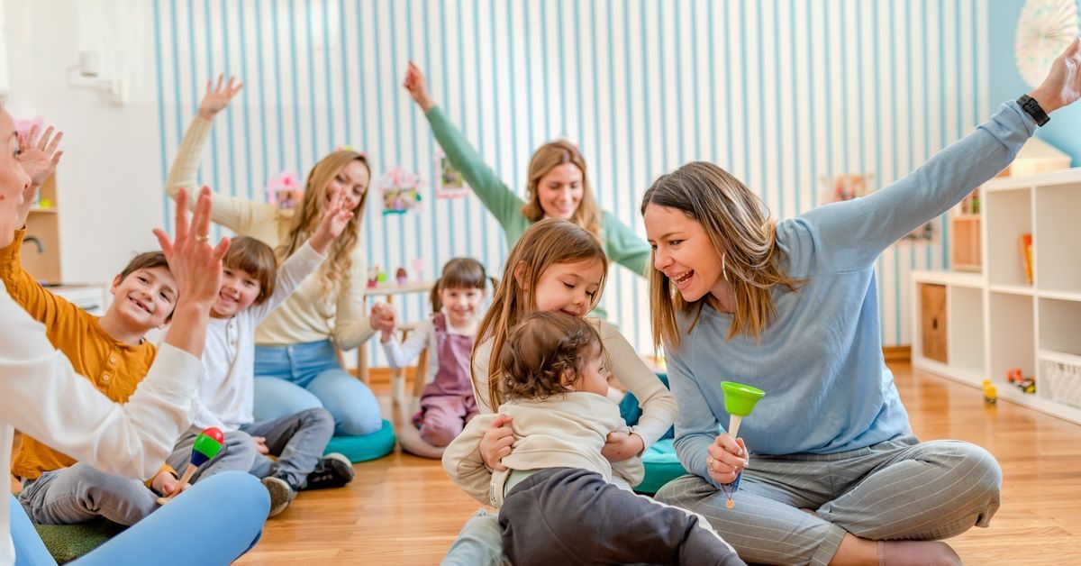 Parents and children sit in a circle cheering as an instructor leads the group on a bright classroom floor.