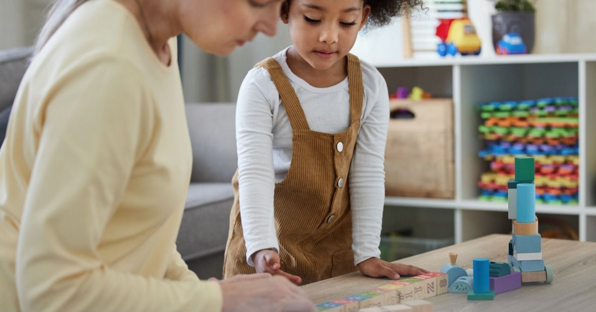 A therapist and young girl stack colorful wooden blocks together at a table in a bright ABA therapy room.