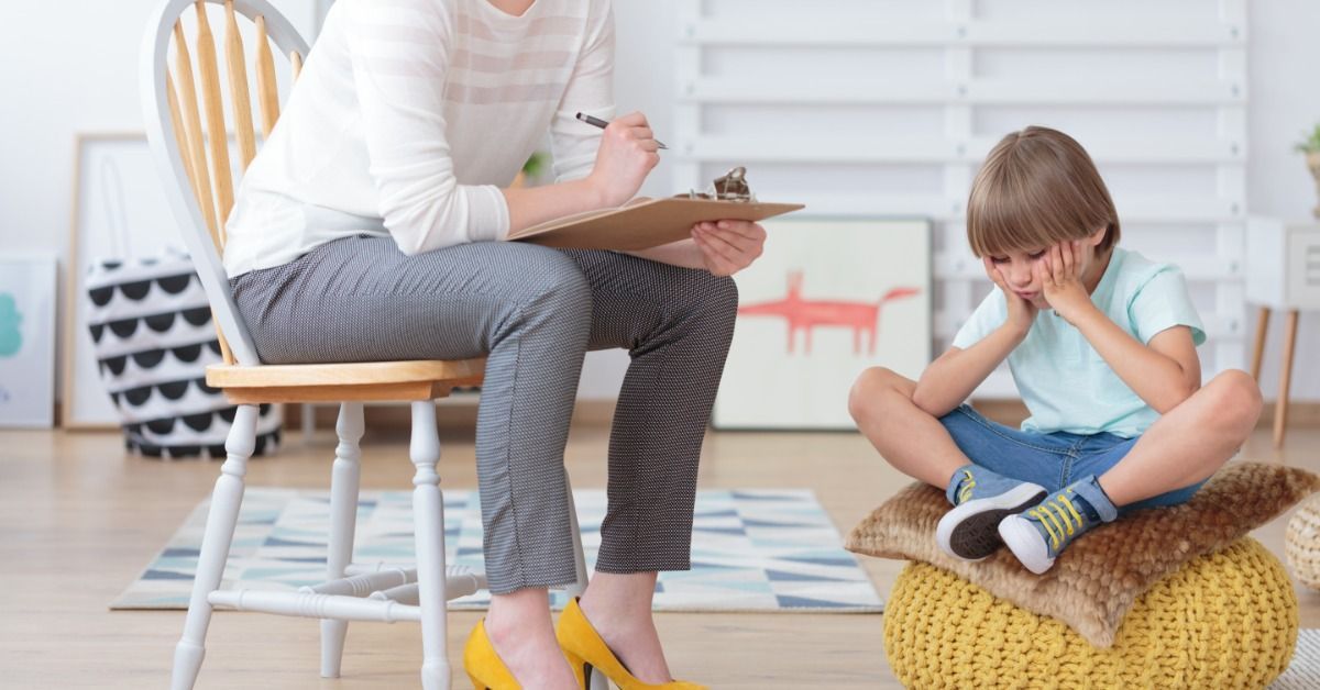 A therapist writes on clipboard while a young boy sits on a cushion with his hands on his cheeks in a bright therapy room.