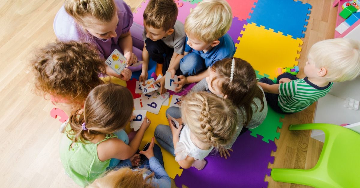 A teacher sits with young children in a circle on colorful mats during a group learning activity.
