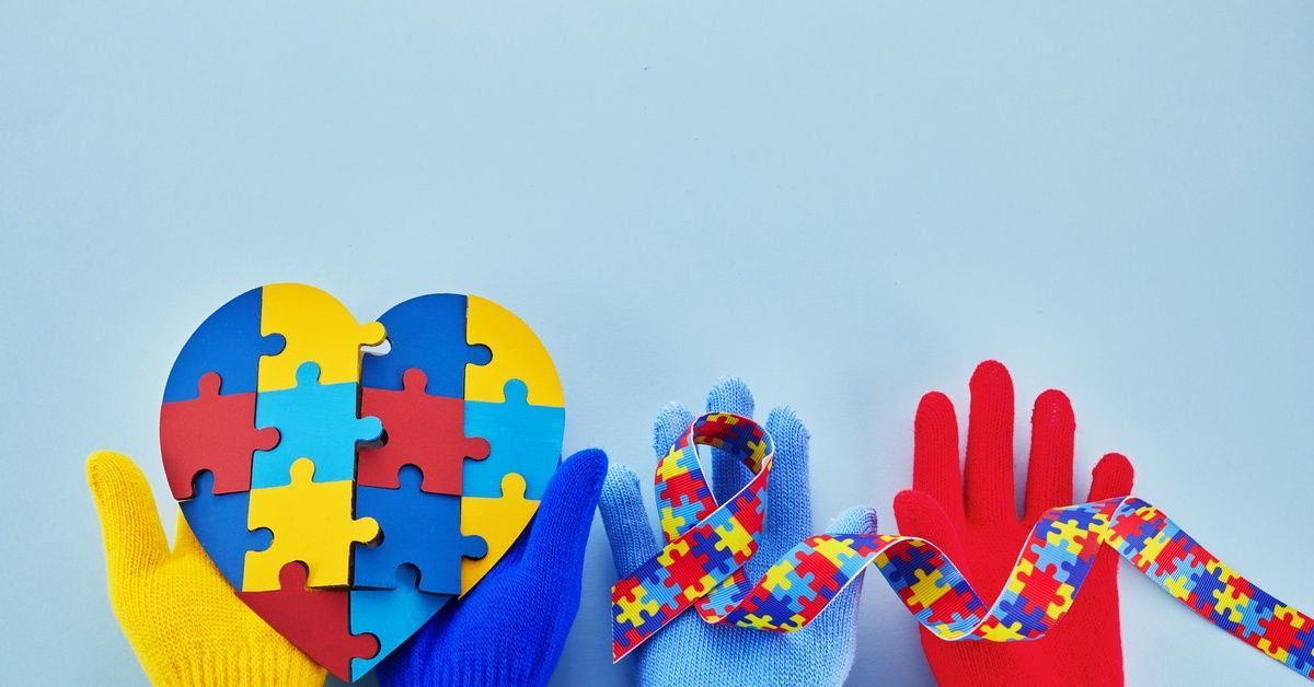 Colorful gloved hands hold a heart-shaped puzzle and an autism awareness ribbon.