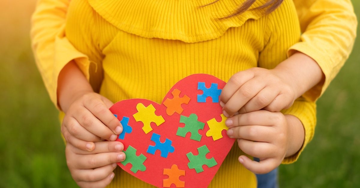 Two children in yellow shirts hold a red heart covered with colorful puzzle pieces against a grassy background