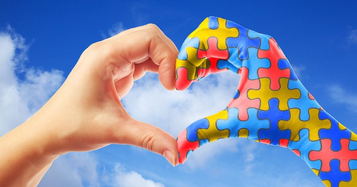 A Caucasian hand and a puzzle-painted hand form a heart shape against a bright blue sky with white clouds.
