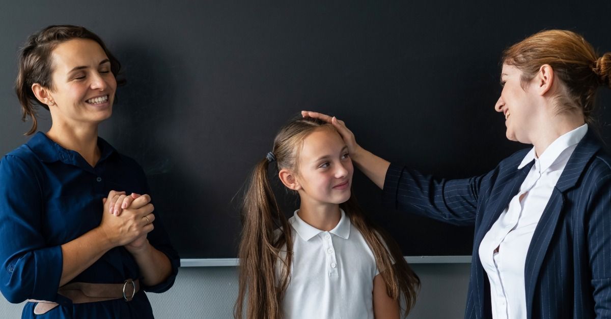A teacher gently pats a smiling schoolgirl’s head while a mother stands beside them.