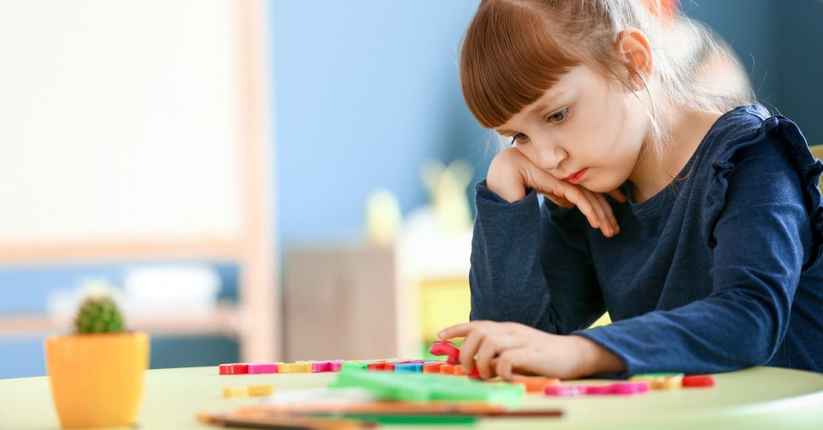 A young girl rests her chin on her hand while arranging colorful blocks on a table in a bright classroom.