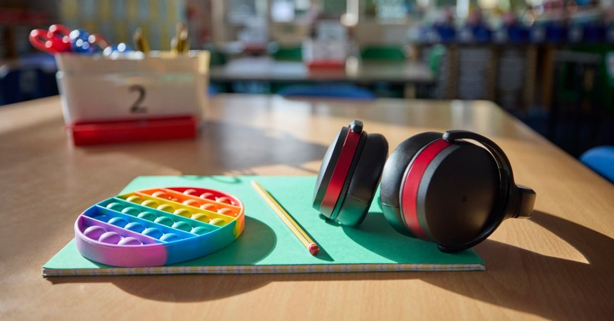 A pair of black headphones, a pencil, and a rainbow pop-it toy sit on a notebook resting on a classroom desk.