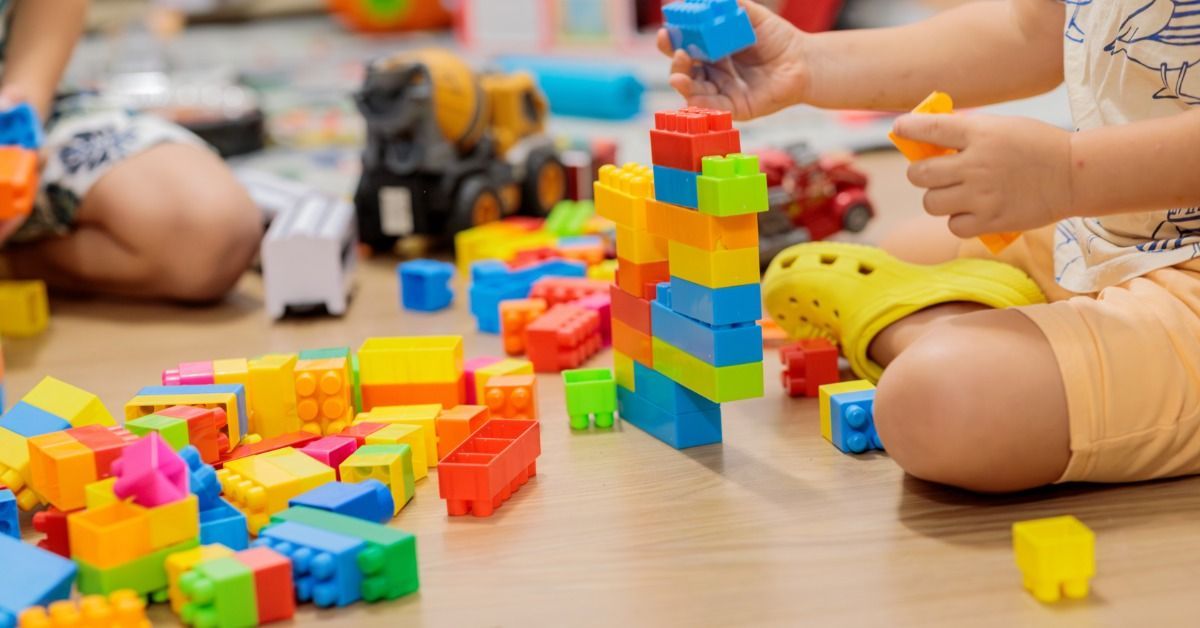 A child places a blue block on a colorful tower while sitting on the floor surrounded by colorful building blocks.