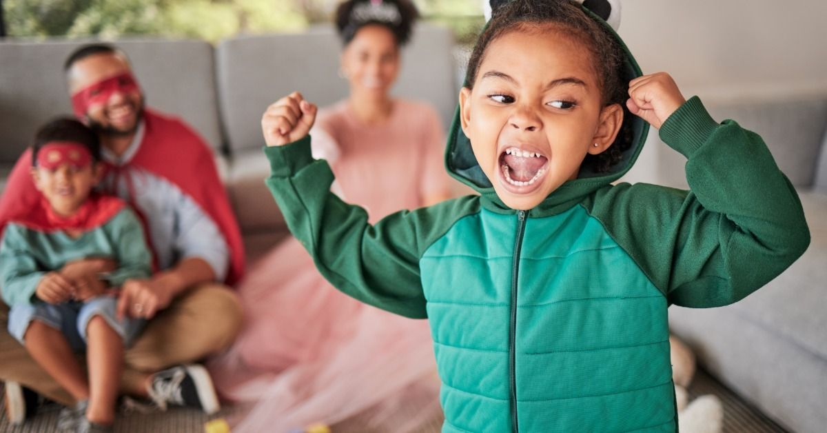 A young child in a green dinosaur costume raises their arms and roars while their family watches and plays in the background.