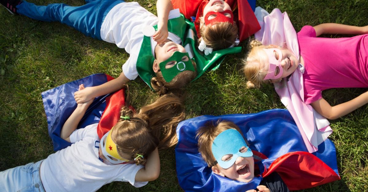 Children wearing colorful superhero capes and masks lie in a circle on the grass.