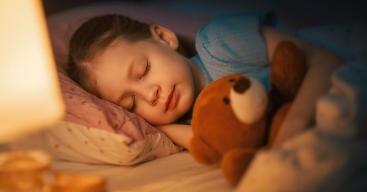 A young child sleeps peacefully in bed while cuddling a brown teddy bear, illuminated by a soft bedside lamp.