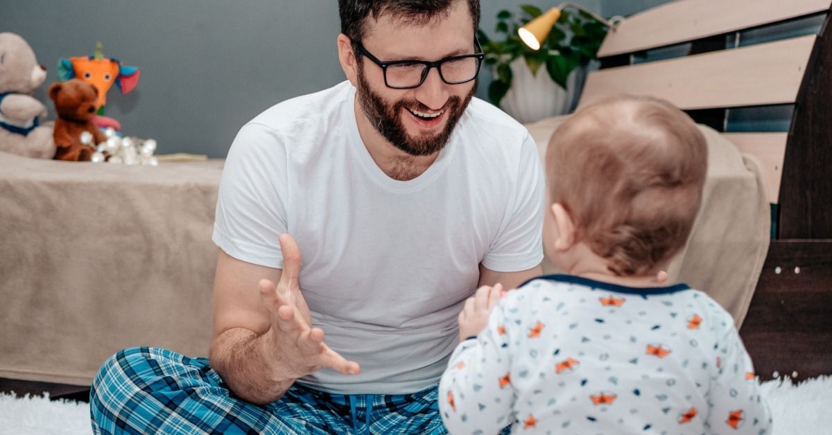A smiling father in glasses and pajamas sits on the floor near his baby. The baby sits on a potty training toilet.