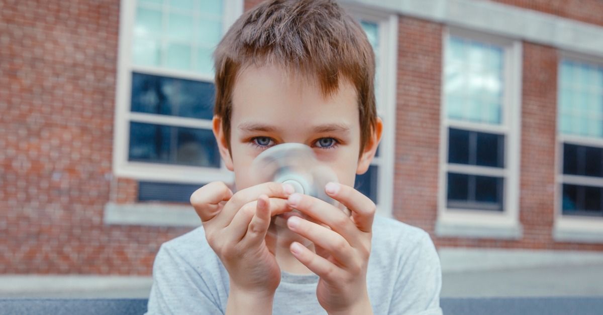 A young boy holds a white fidget spinner with both hands while standing outside in front of a brick building.