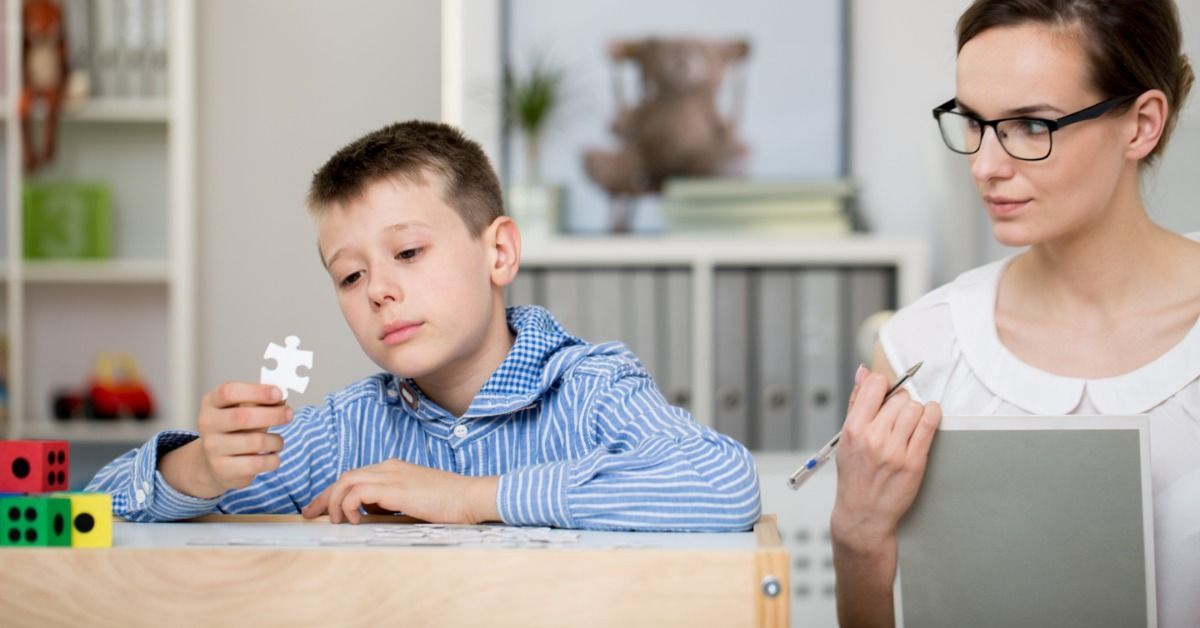 A young boy examines a white puzzle piece at a table while an ABA therapist with glasses observes him.