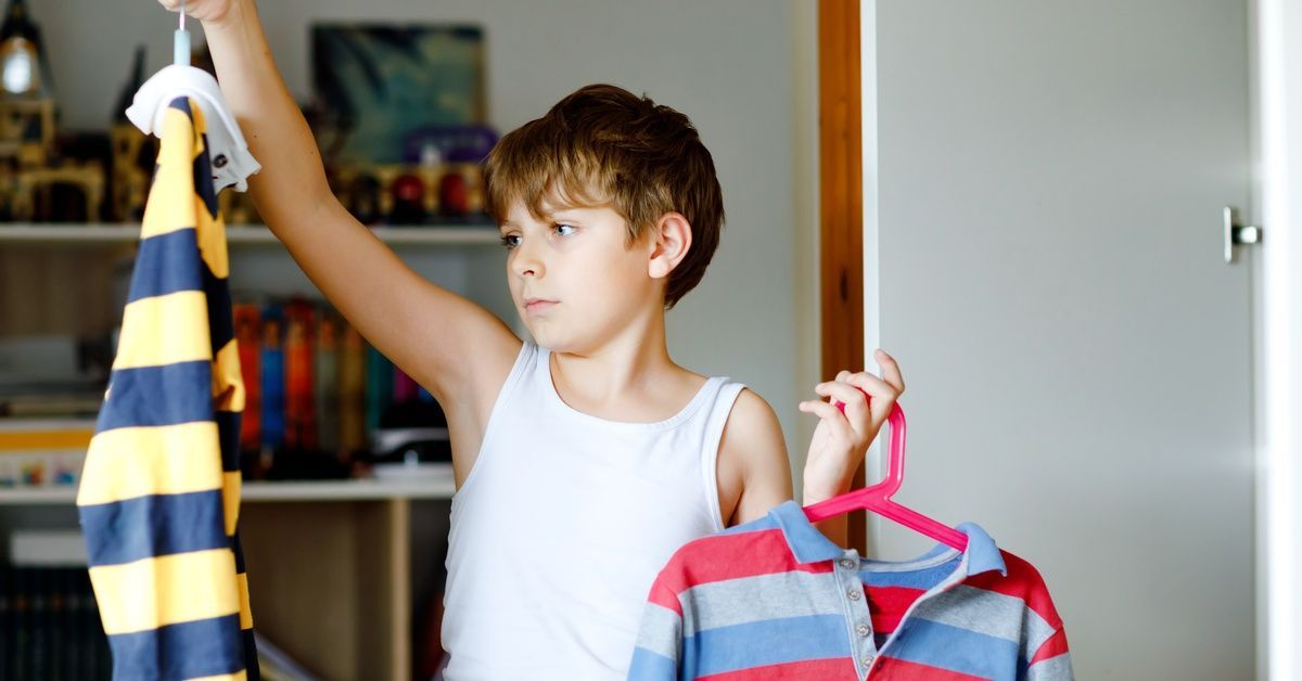 A young boy holds two striped shirts on hangers while standing in a bedroom and getting dressed for school.