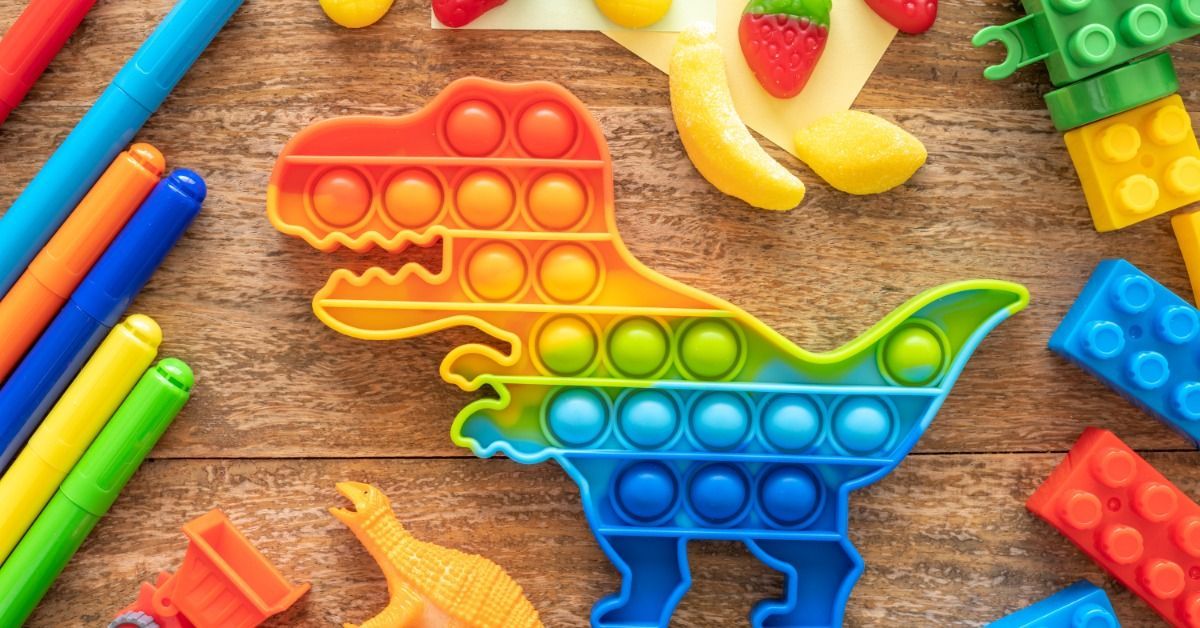 A colorful dinosaur-shaped pop-it toy sits on a table surrounded by markers, toy blocks, and small fruit-shaped fidgets.
