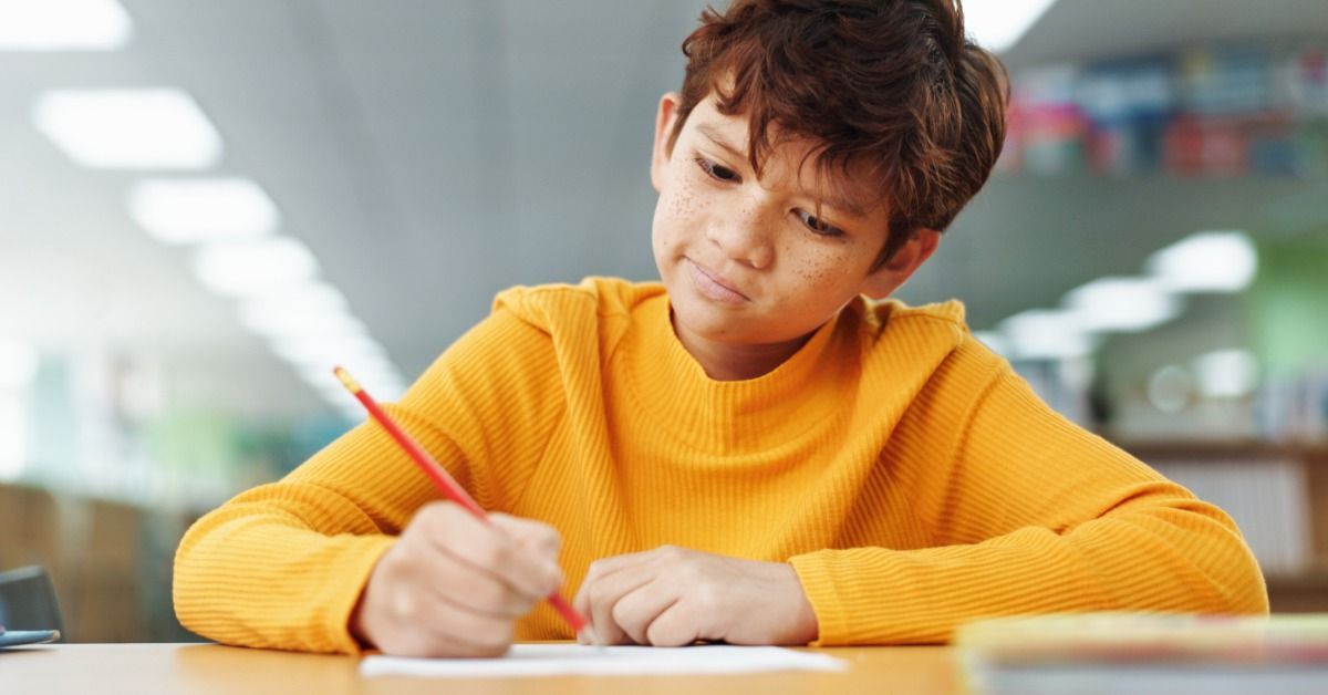 A young boy wearing a yellow sweater and holding a red pencil finishes his test in the school library.
