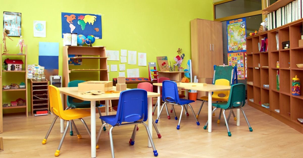 A classroom contains chairs around small tables, with shelves of toys and books along the wall.