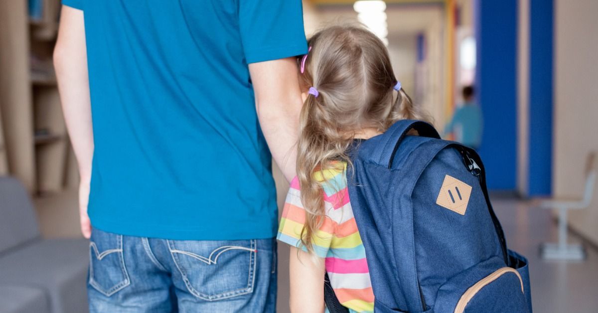 An parent walks their child with a backpack down a school hallway, as the child holds onto the adult’s arm for support.