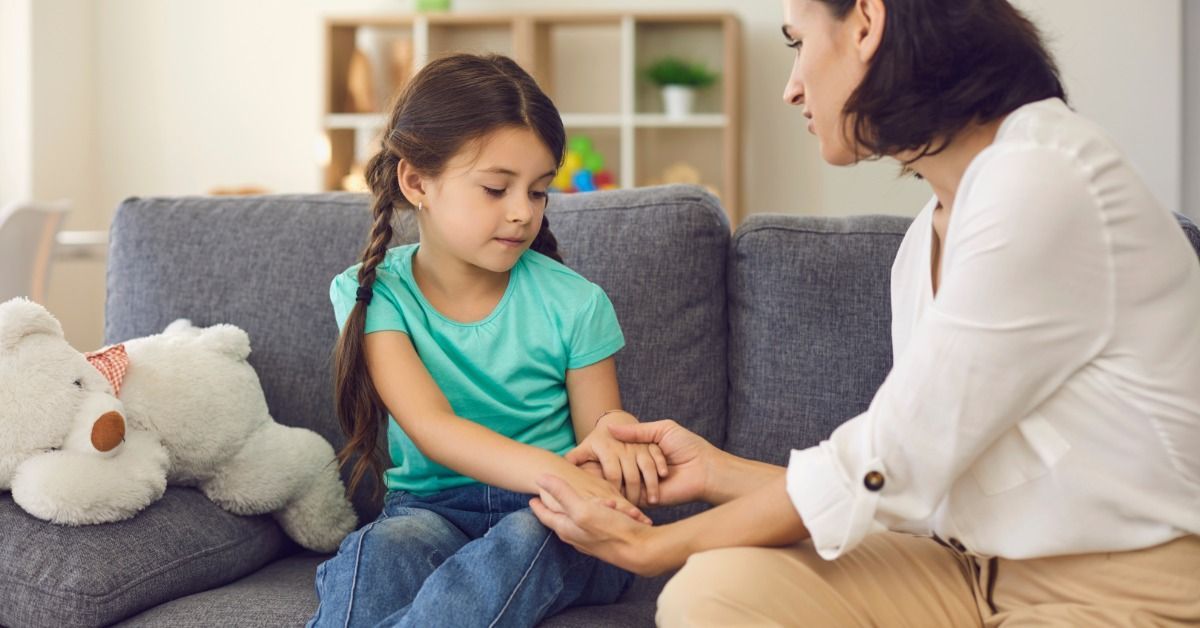 A parent gently holds their daughter's hands on a couch while talking. A stuffed bear lays next to the daughter.