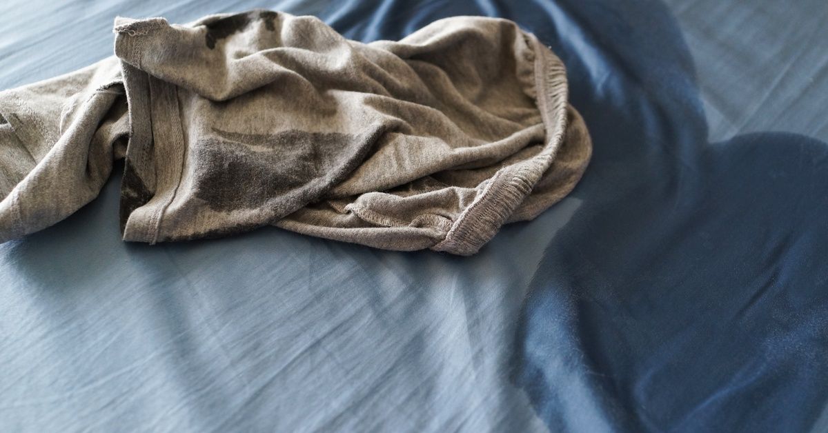 A damp stain spreads across a bed sheet while a pair of wet pants rests on top, showing a recent nig