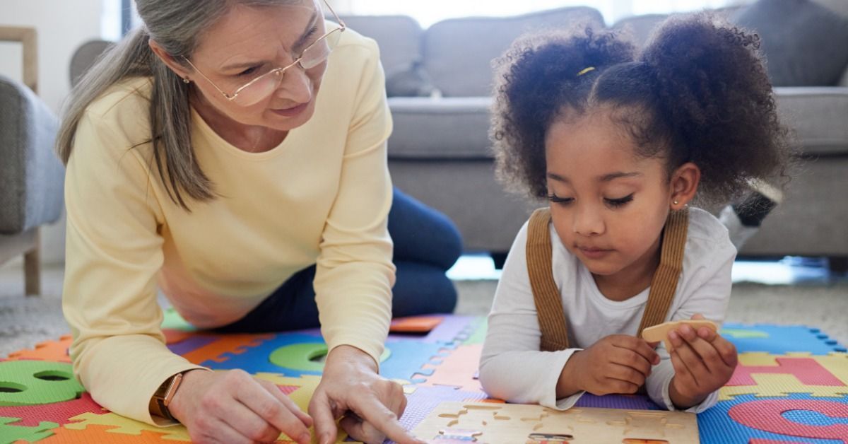 A therapist helps a young girl complete a wooden puzzle on a colorful foam mat in a living room