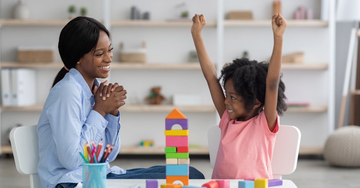 A woman watches a girl raise her arms in excitement after stacking colorful blocks on a table.