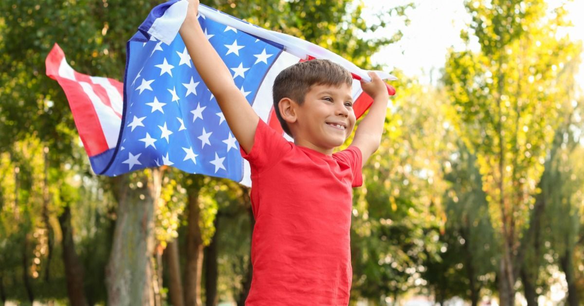 A smiling boy holds an American flag over his shoulders in a sunny park with green trees behind him.