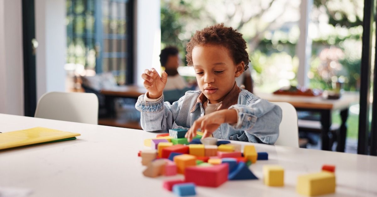 A young boy stacks colorful blocks on a table in a bright room while focusing on arranging shapes.