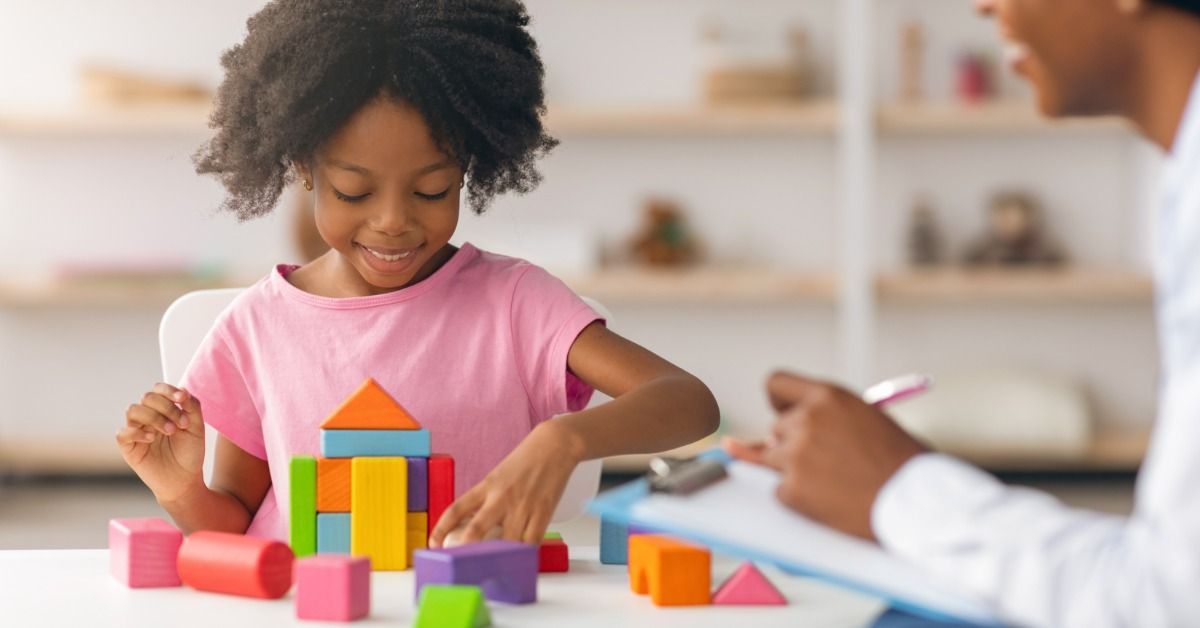 A smiling girl builds a colorful block tower while a clinician takes notes beside her.