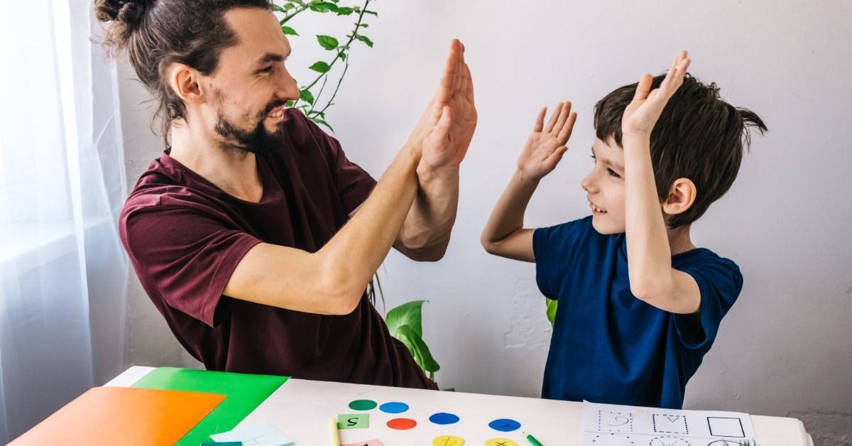 A therapist and a young boy raise their hands for a high five during a playful learning activity at a table.