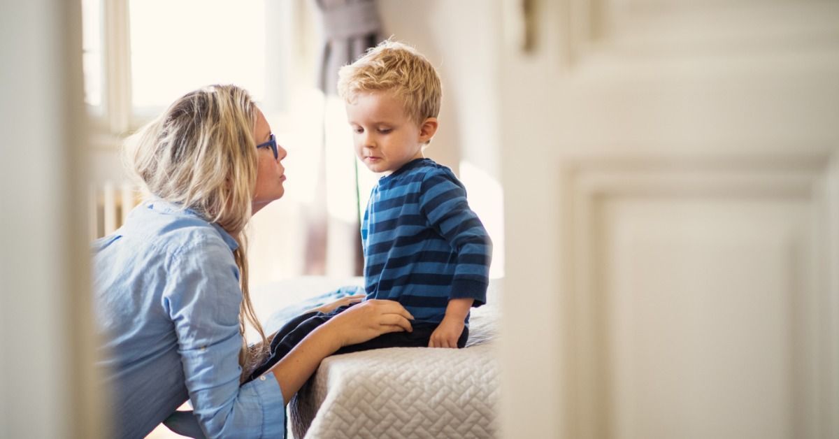 A mother kneels beside her young child on a bed and talks with him calmly in a softly lit bedroom at home.