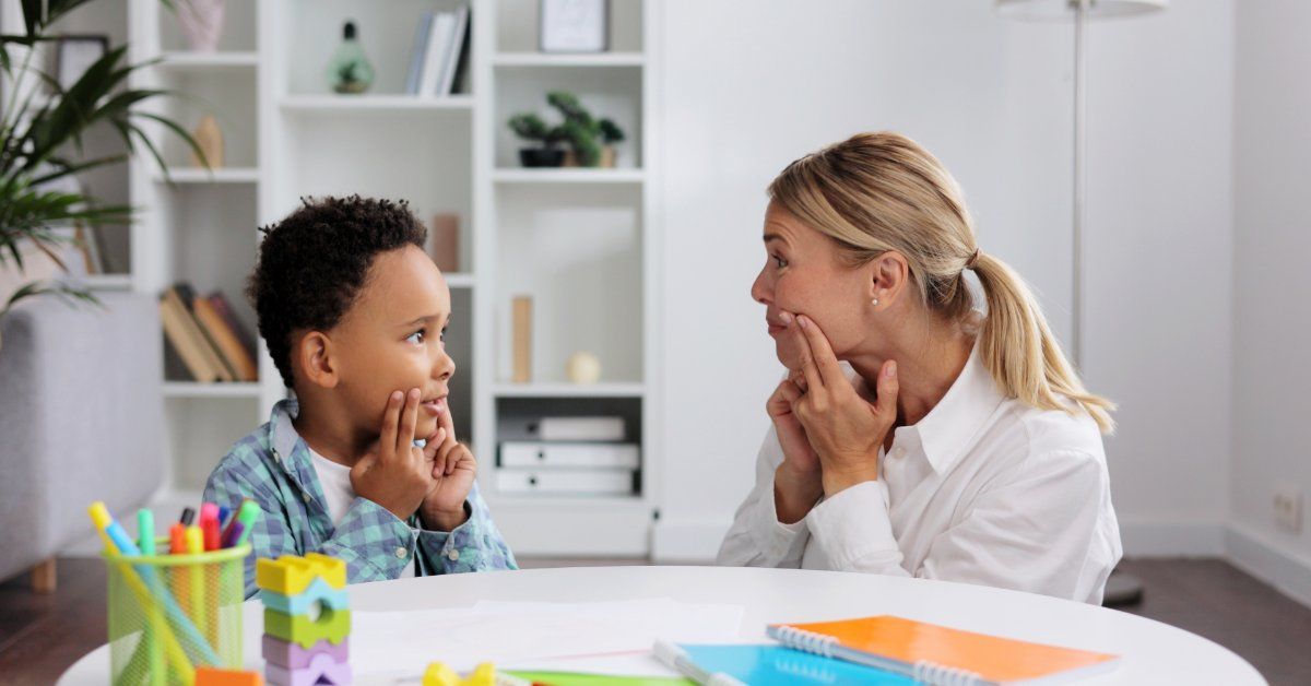 A therapist and a young boy mirror face-touching gestures at a table with colorful learning toys.