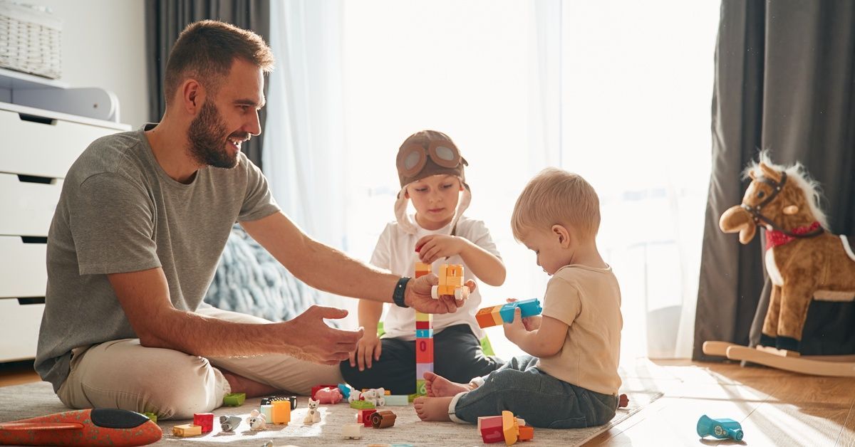A father and two young children build a block tower together on a sunlit playroom floor with a toy horse nearby.