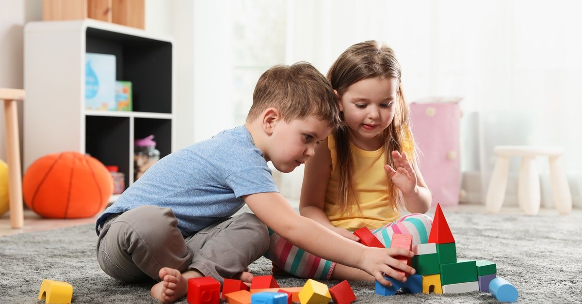 A boy and a girl build a colorful block tower together on a soft rug in a bright playroom with scattered toys.