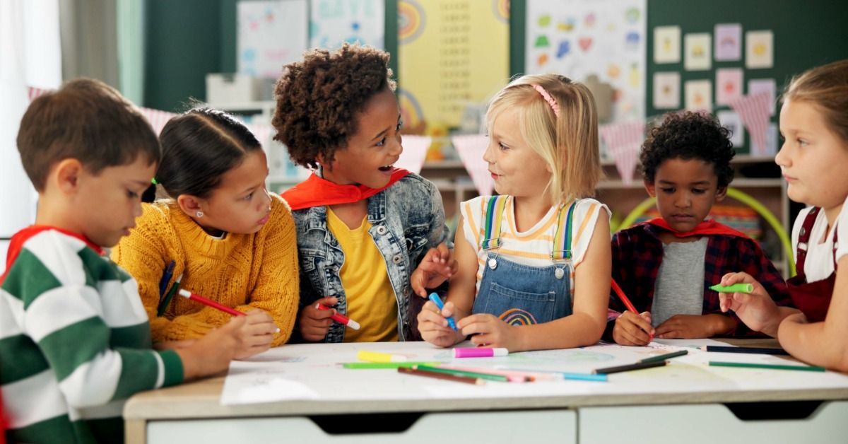 Children sit around a classroom table and draw with markers while two girls talk during a group activity.
