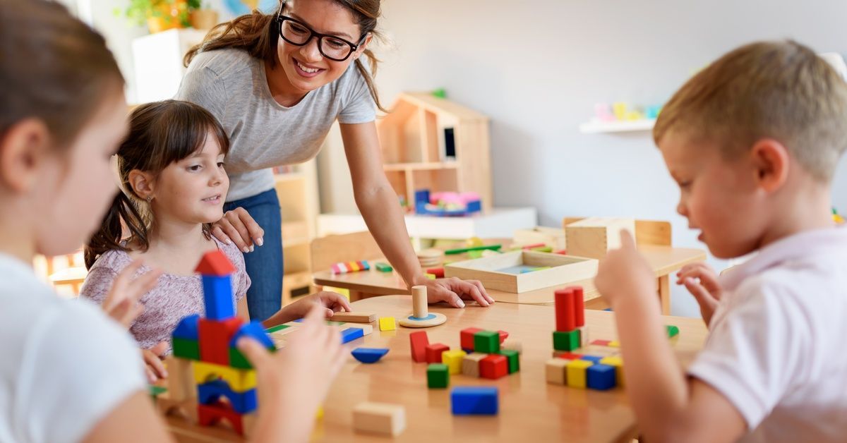 A smiling teacher guides young children as they build structures with colorful wooden blocks at a table.
