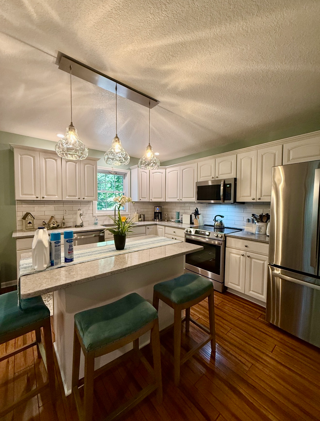 Bright white kitchen with granite island, green stools, pendant lights, and stainless steel appliances.