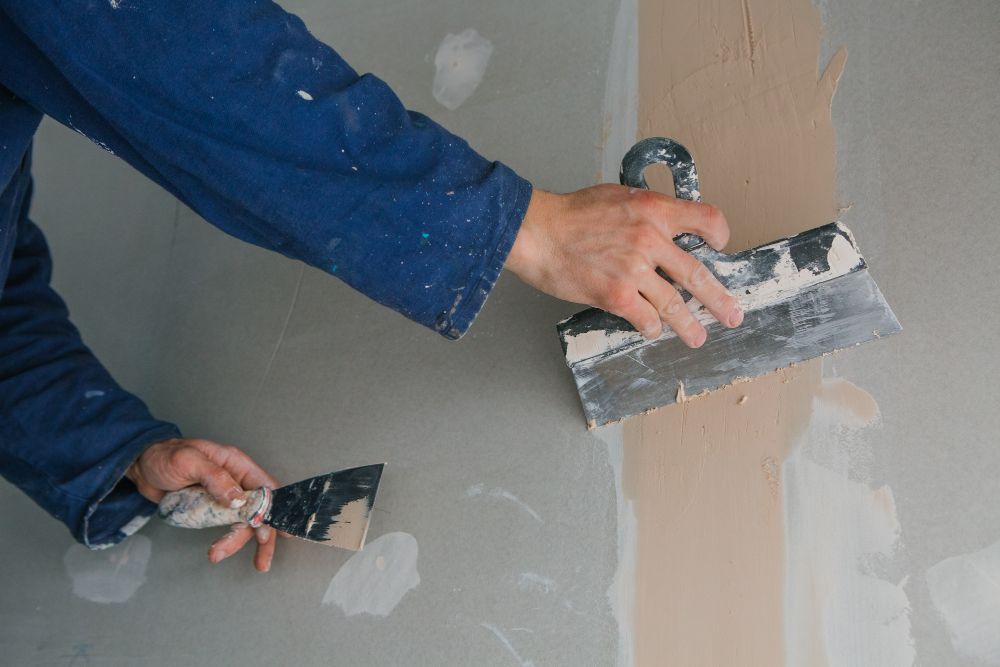 Person in blue coveralls using a putty knife to apply plaster to a wall.