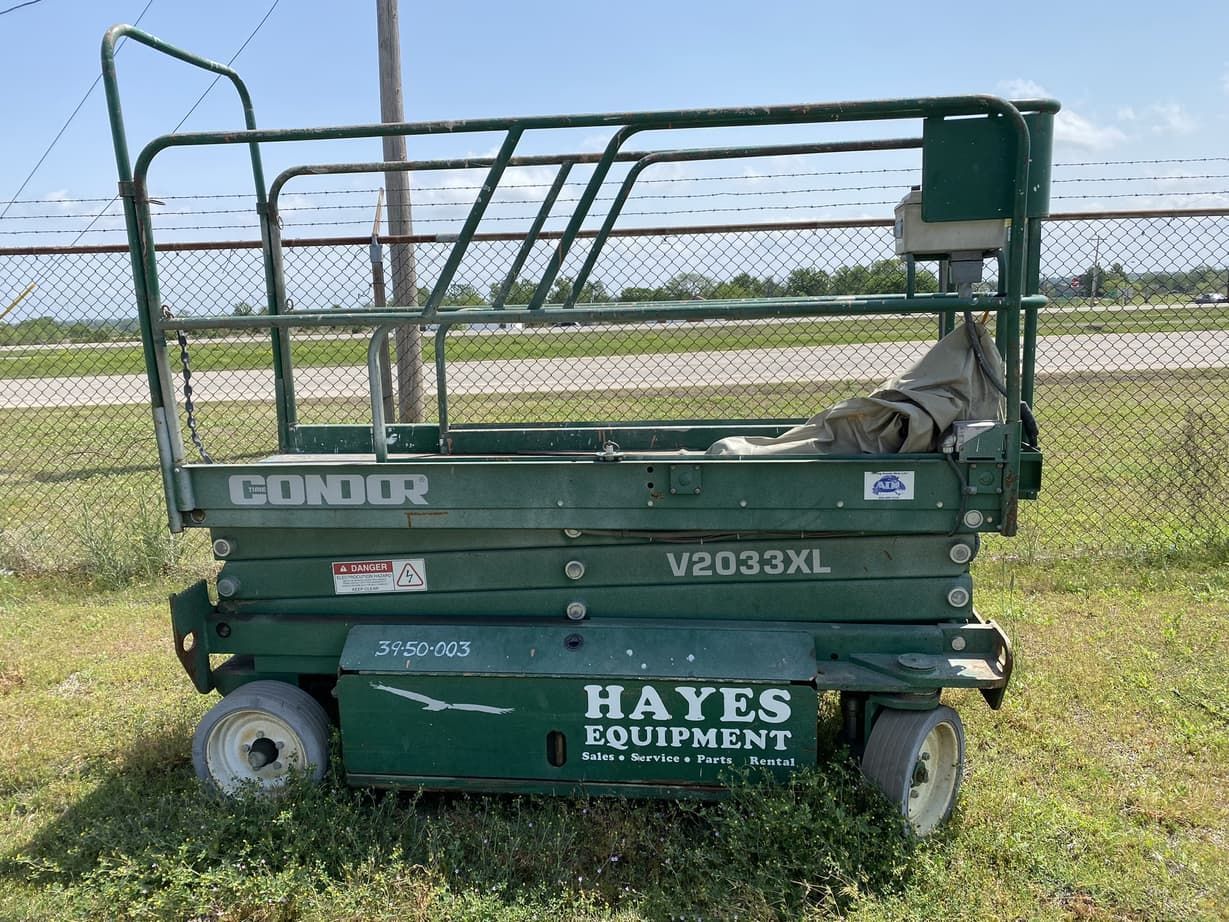 A green scissor lift is parked in a grassy field next to a chain link fence.