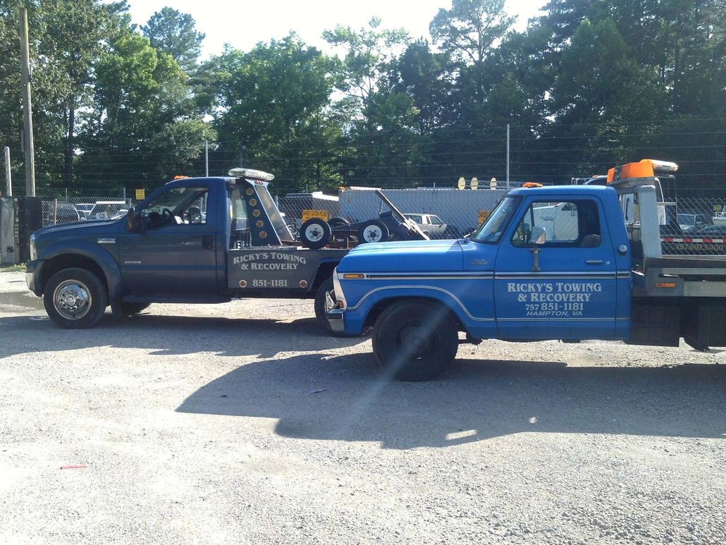 Two blue towing trucks parked side-by-side in a gravel lot on a sunny day.