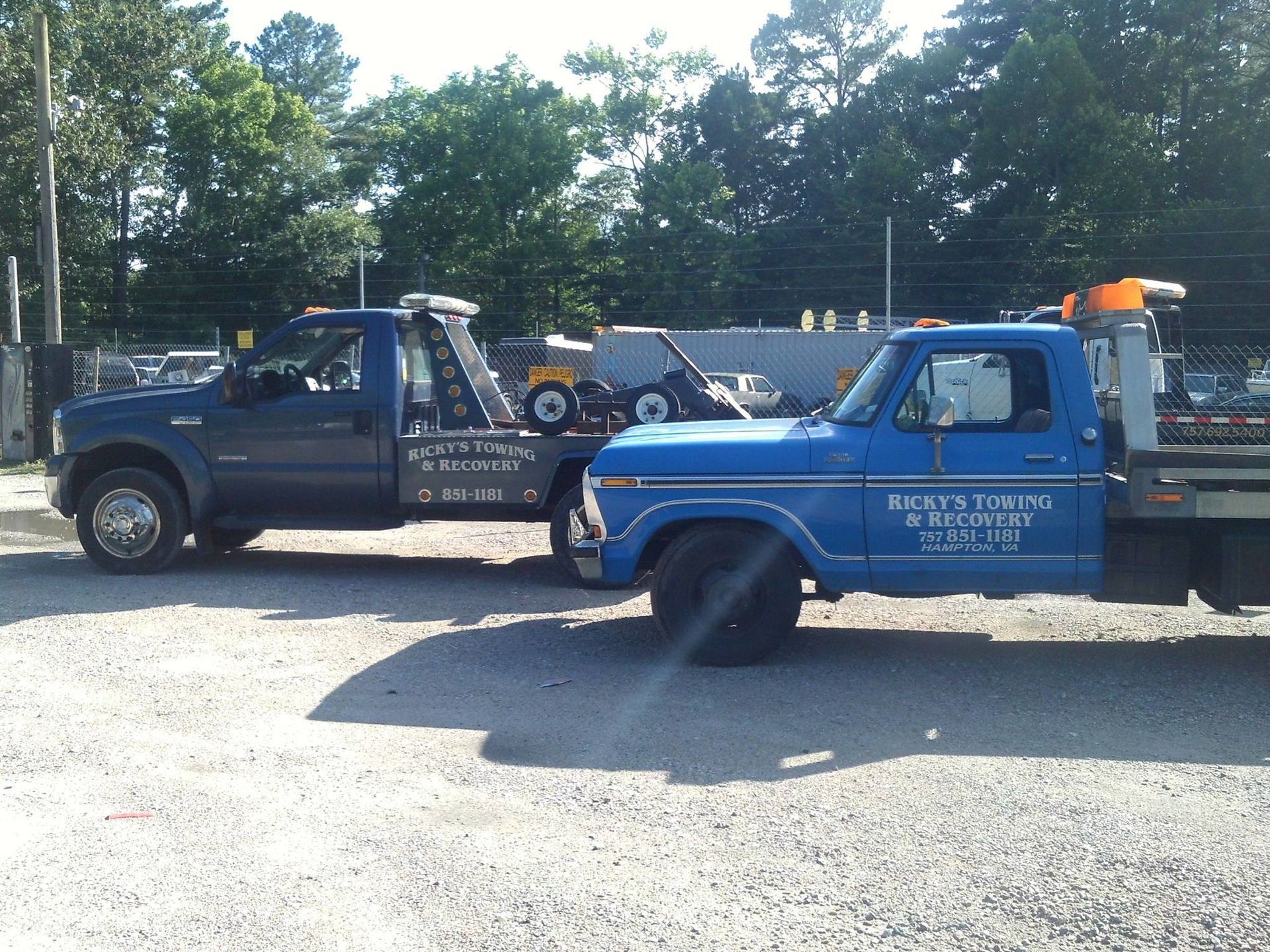 Two blue towing trucks parked side-by-side in a gravel lot on a sunny day.