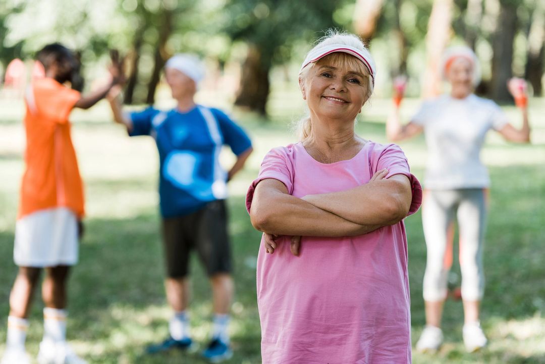 Una donna con una maglietta rosa e una visiera è in piedi con le braccia incrociate in un parco, mentre sullo sfondo si vedono altre tre persone che fanno esercizio fisico.