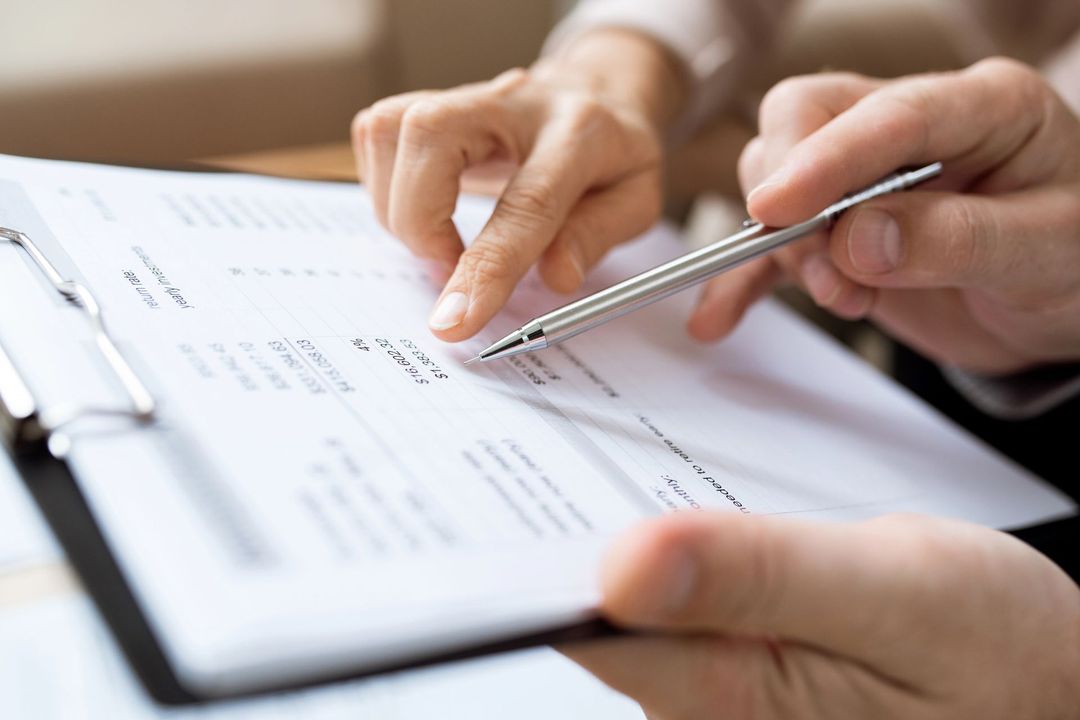 Primo piano di mani che esaminano un documento su un blocco appunti, con una persona che usa una penna argentata per indicare dati specifici.