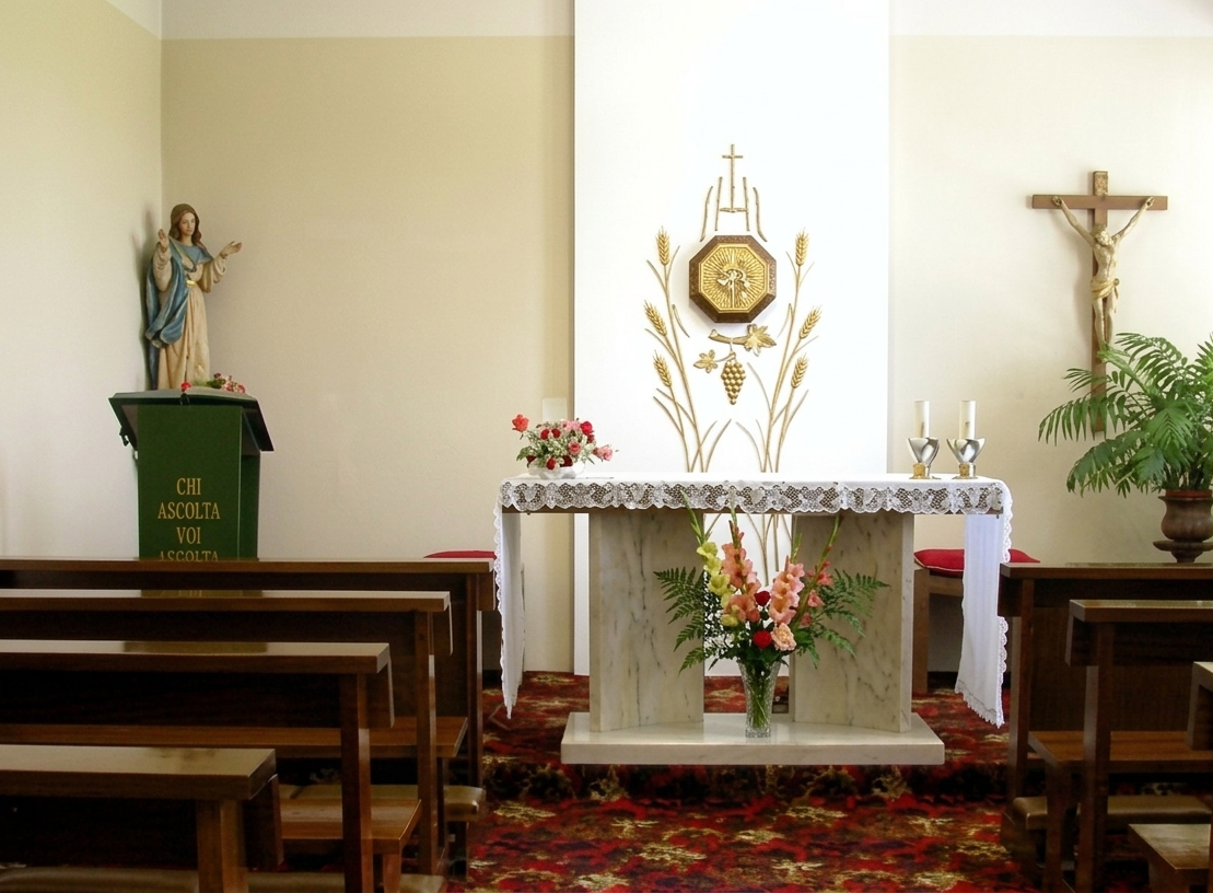 Interno di una piccola cappella con banchi di legno, un altare, un crocifisso e una statua di un santo.