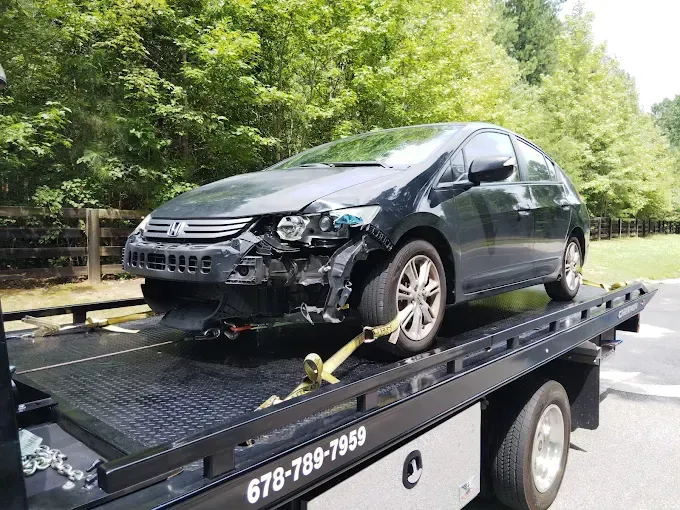 Damaged black car on a tow truck, front end wrecked. Outdoors, sunny day.
