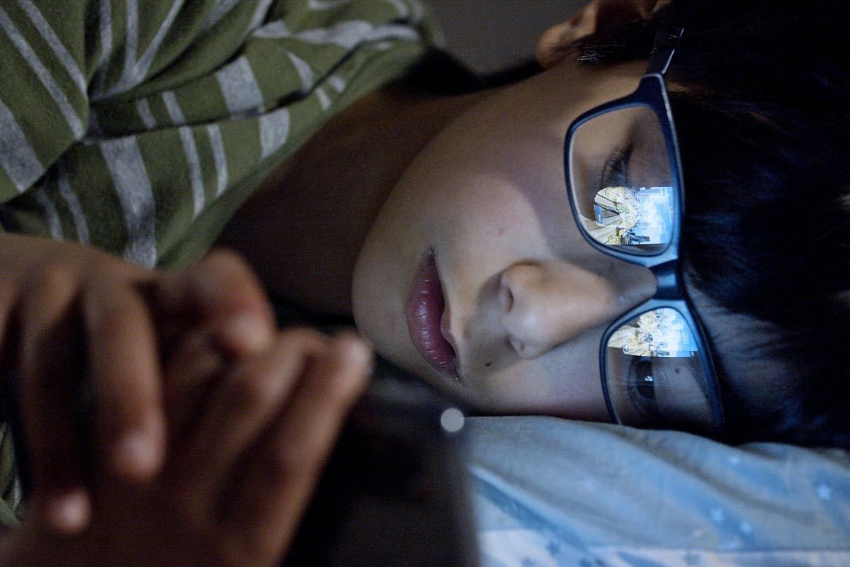 A person lying in bed at night, illuminated by the glow of a smartphone screen reflecting in their eyeglasses.