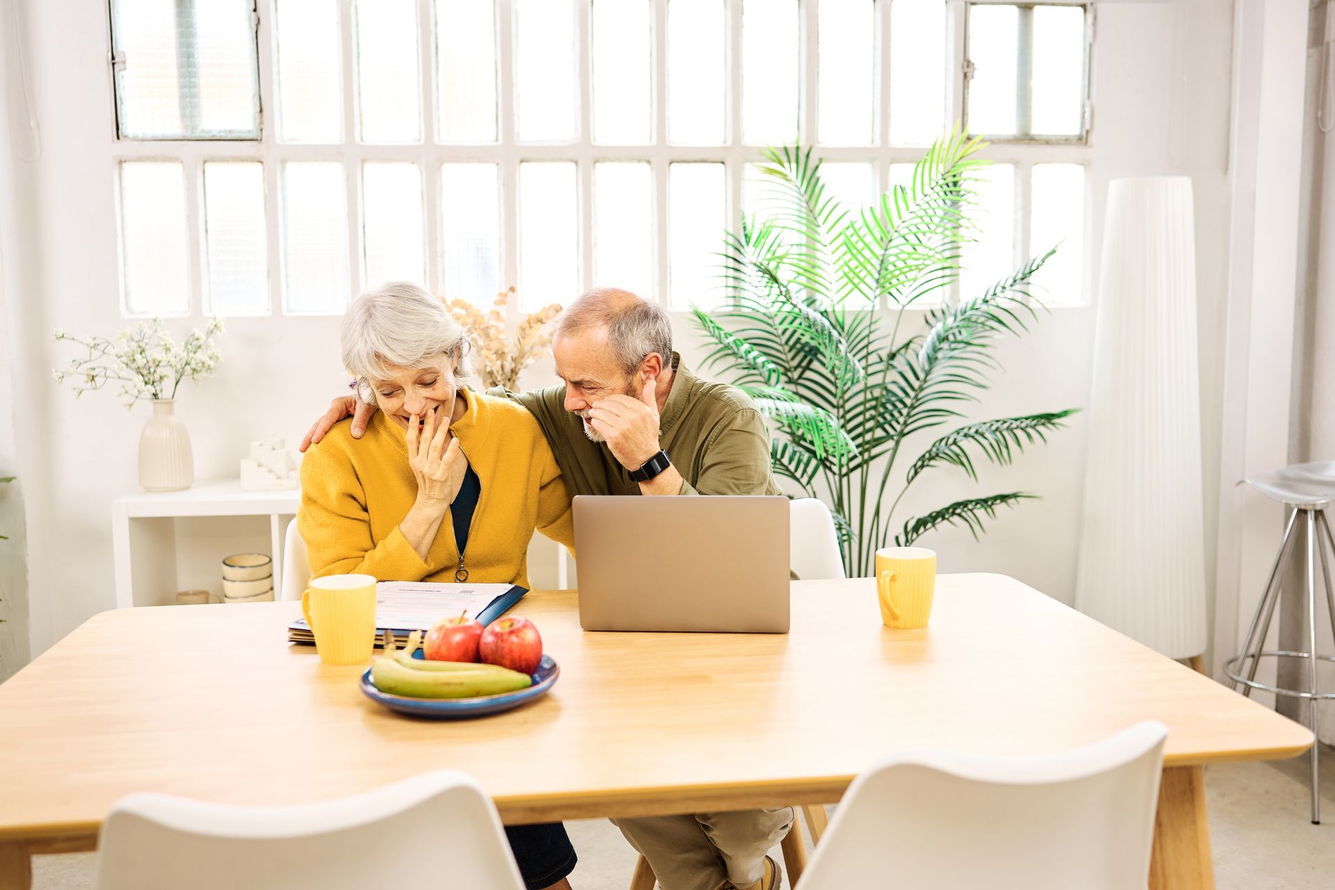 An elderly couple is sitting at a table looking at a laptop.