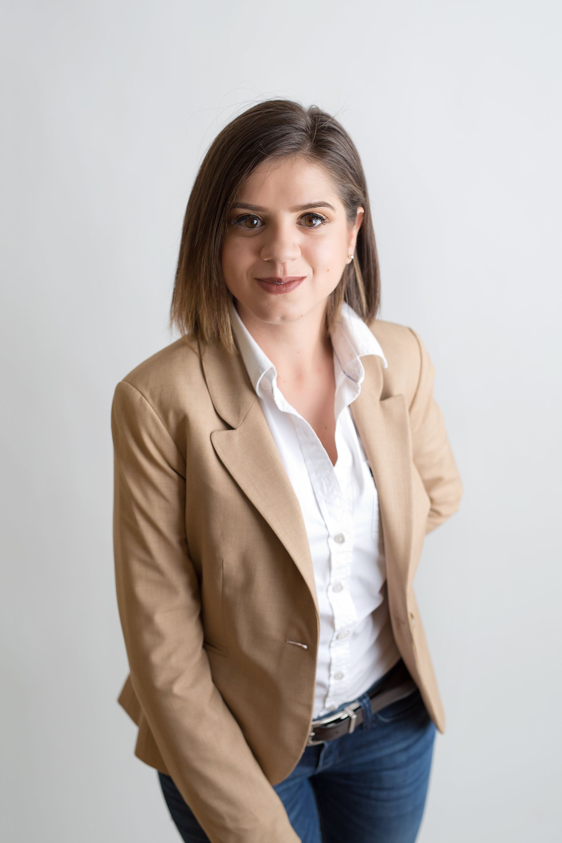 A woman in a tan jacket and white shirt is standing in front of a white wall.